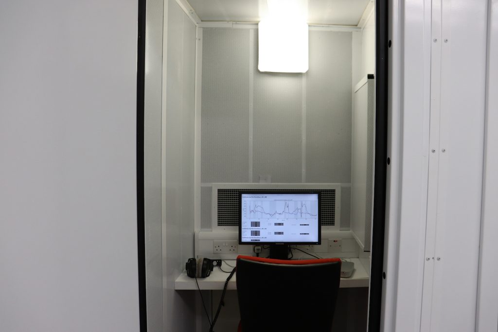 Image shows a small enclosed space with a desktop computer, headphones and chair