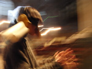 A person in a blurry image with lights behind them. The way the camera has caught the light makes the image appear to vibrate.
